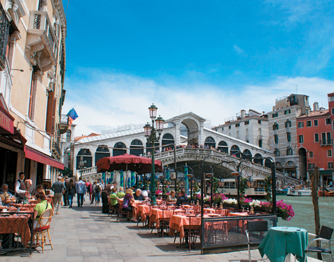 Venice Italy Restaurant-goers in Venice have lunch outdoors with views of canal boats cruising under the Rialto Bridge.